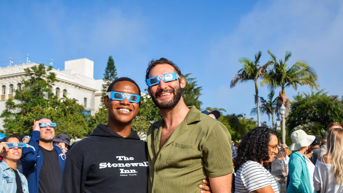 Two men wearing eclipse sunglasses smiling in a large crowd