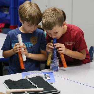 Two young boys holding painted tubes leaning over a beaker of water in a classroom