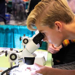 Teenaged blonde boy looks through a microscope on a table
