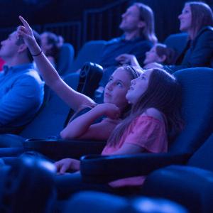Two girls in a dome IMAX theater lean into each other and smile while looking at the screen. The farther girl is pointing up at the dome. 