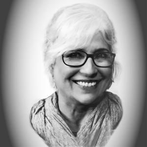 A black and white photo of a middle aged white woman. She is smiling, wearing glasses, and has a shawl around her shoulders. The image is cropped to just her portrait and the edge of the image fades to a circular grey gradient