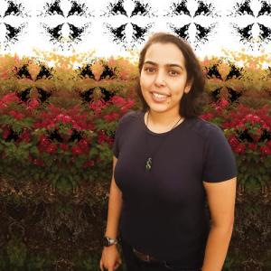 A tan skinned woman with shoulder length dark hair wearing a dark blue tshirt stands in front of a hedge row of green leaves and red flowers. A ink blot style pattern in black is behind her and in front of the hedge.  