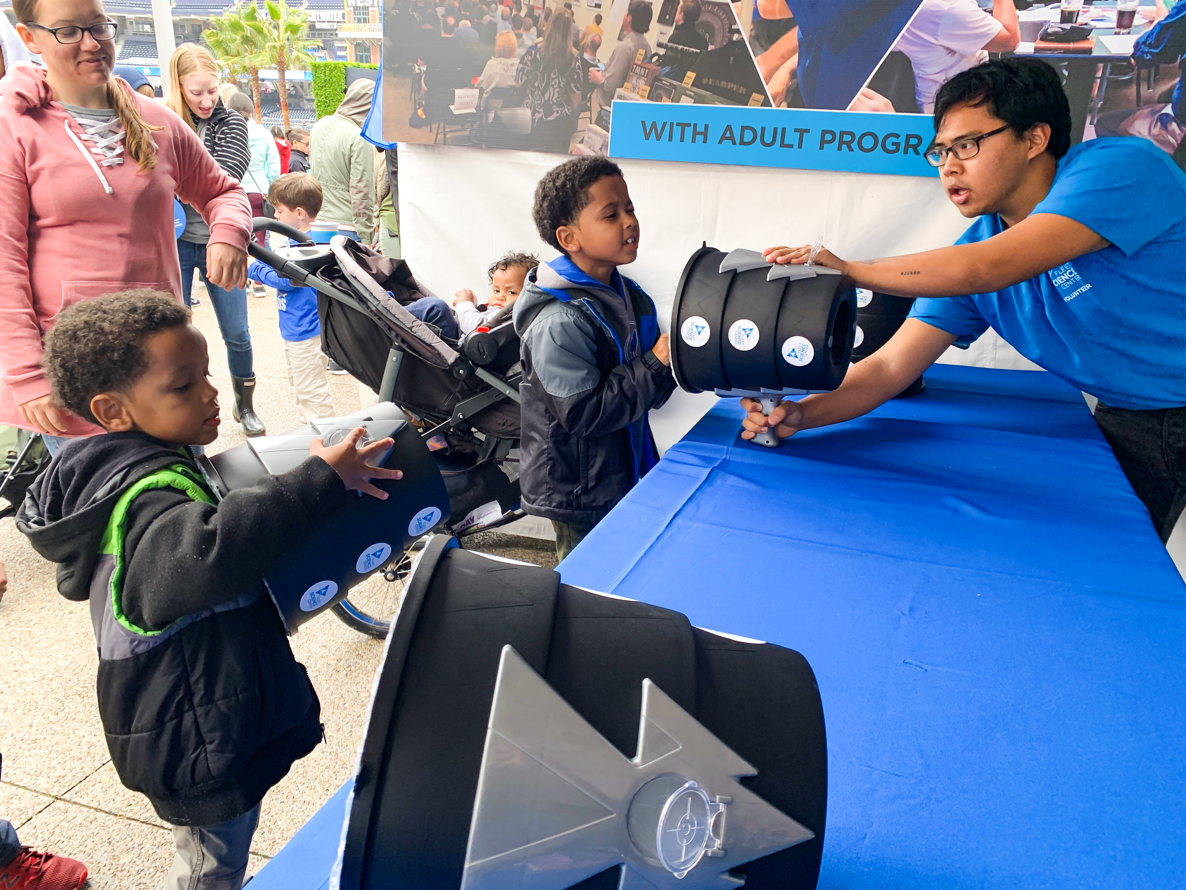 Two young children inspect a container with the assistence of a fleet science center educator at an outdoor community event.