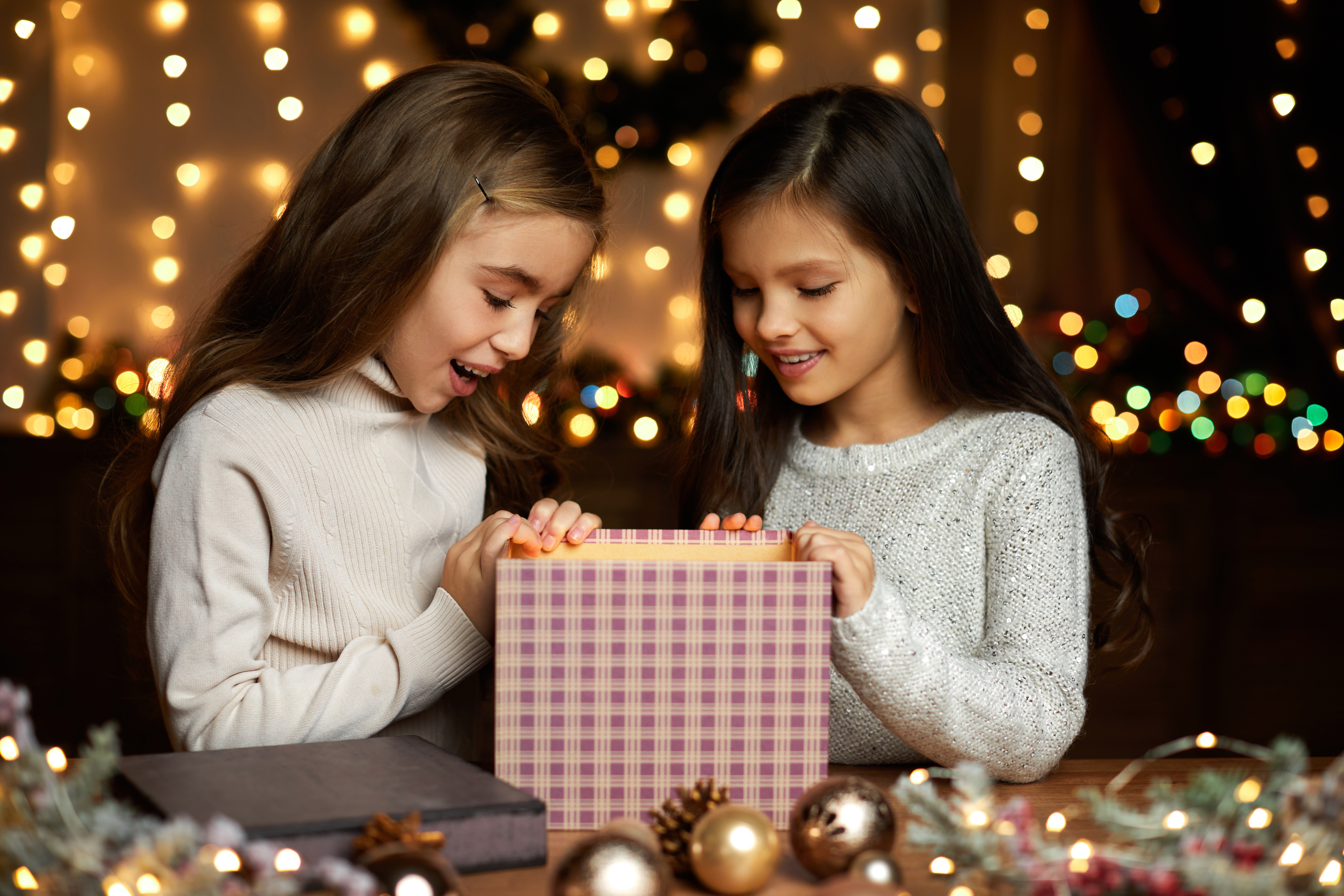 Two young girls open a gift during the holidays surrounded by lights 