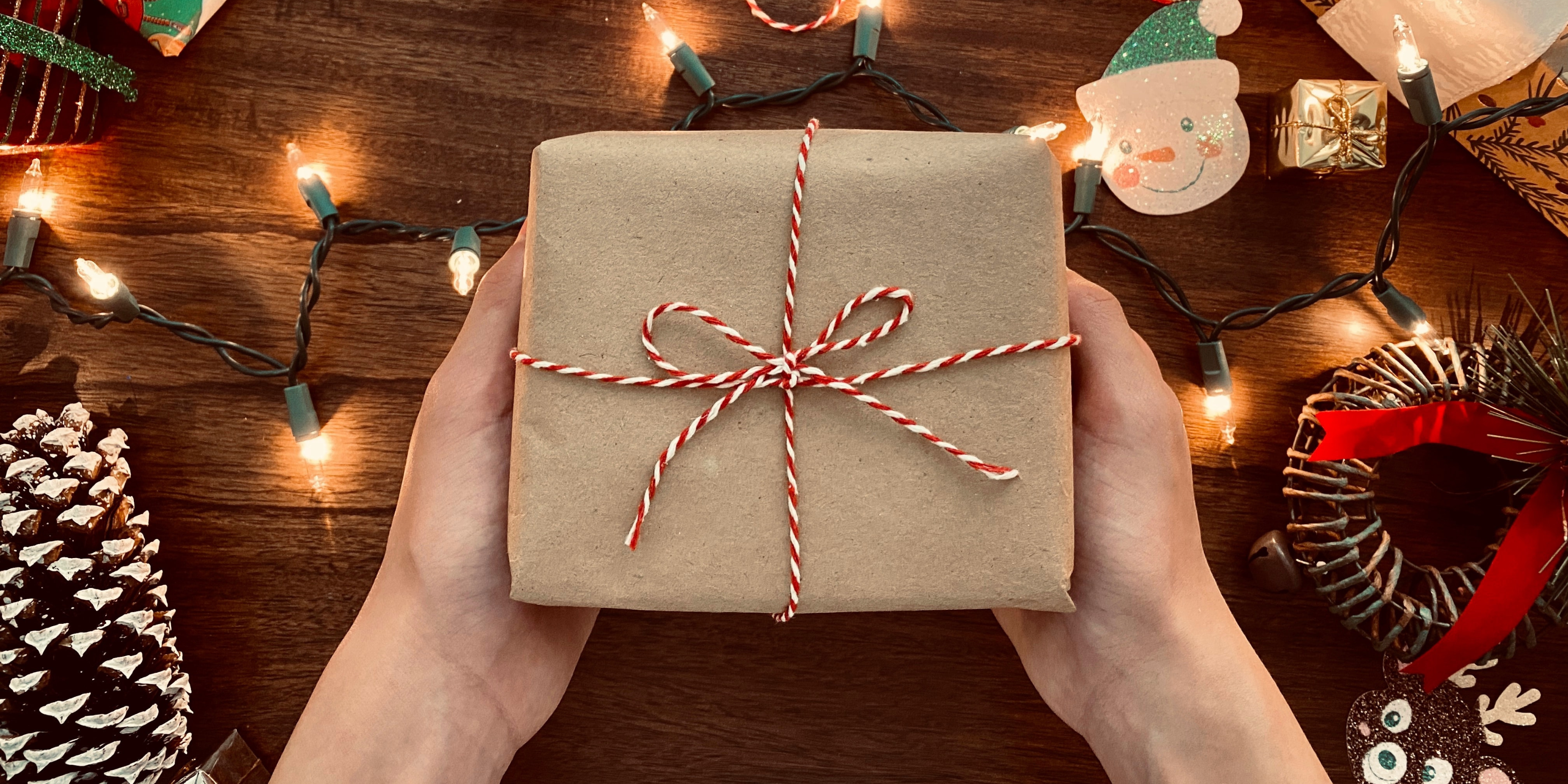 Brown paper wraped gift with a red and white stripped bow being held by a pair of hands with a table and holiday lights in the background.