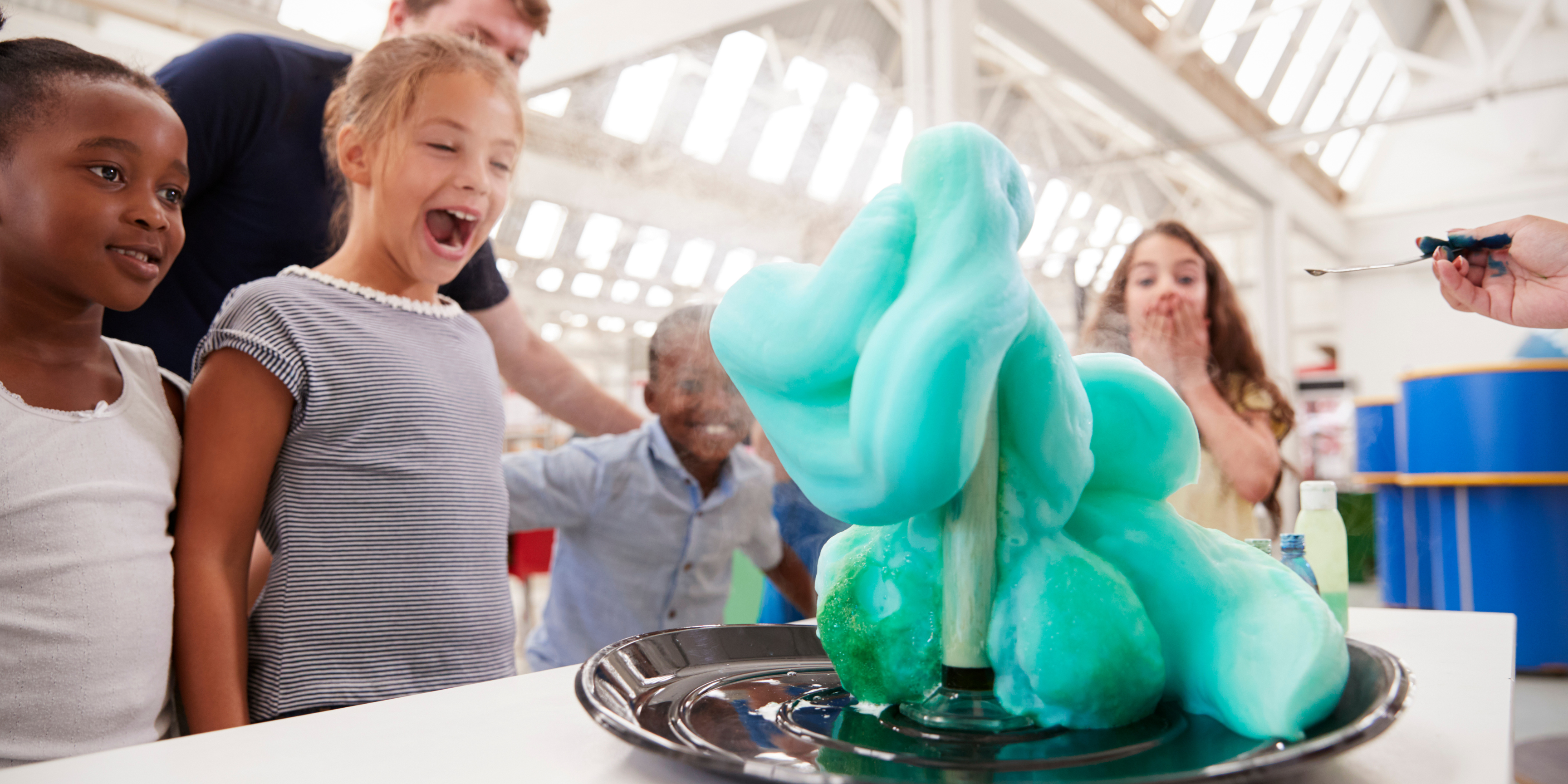 Four children looking at a chemistry science experiment in awe. The experiment is oozing blue and teal foam onto a table.