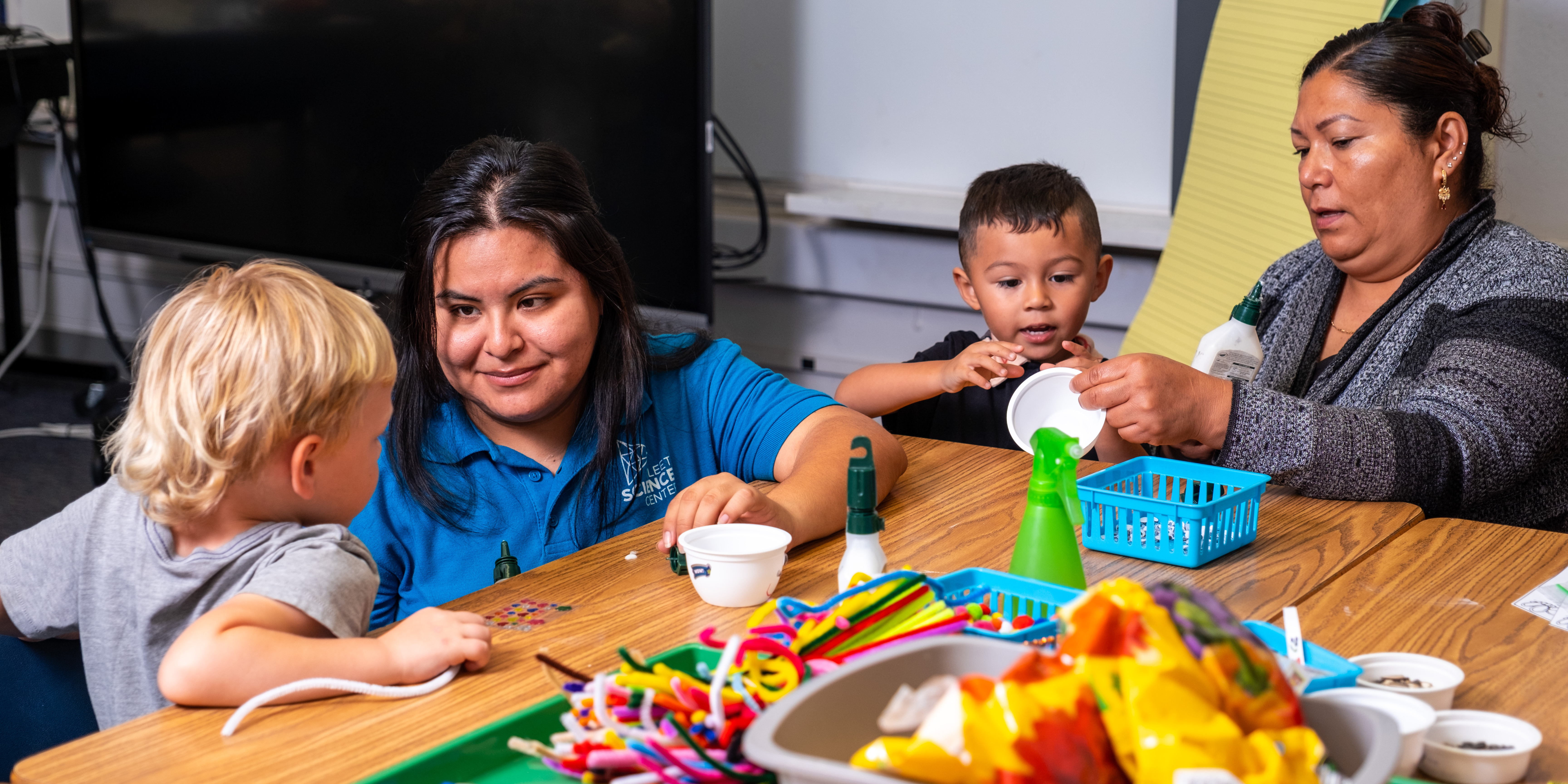 Fleet educator and parent engaging in hands-on STEM learning with two young students inside of a classroom.