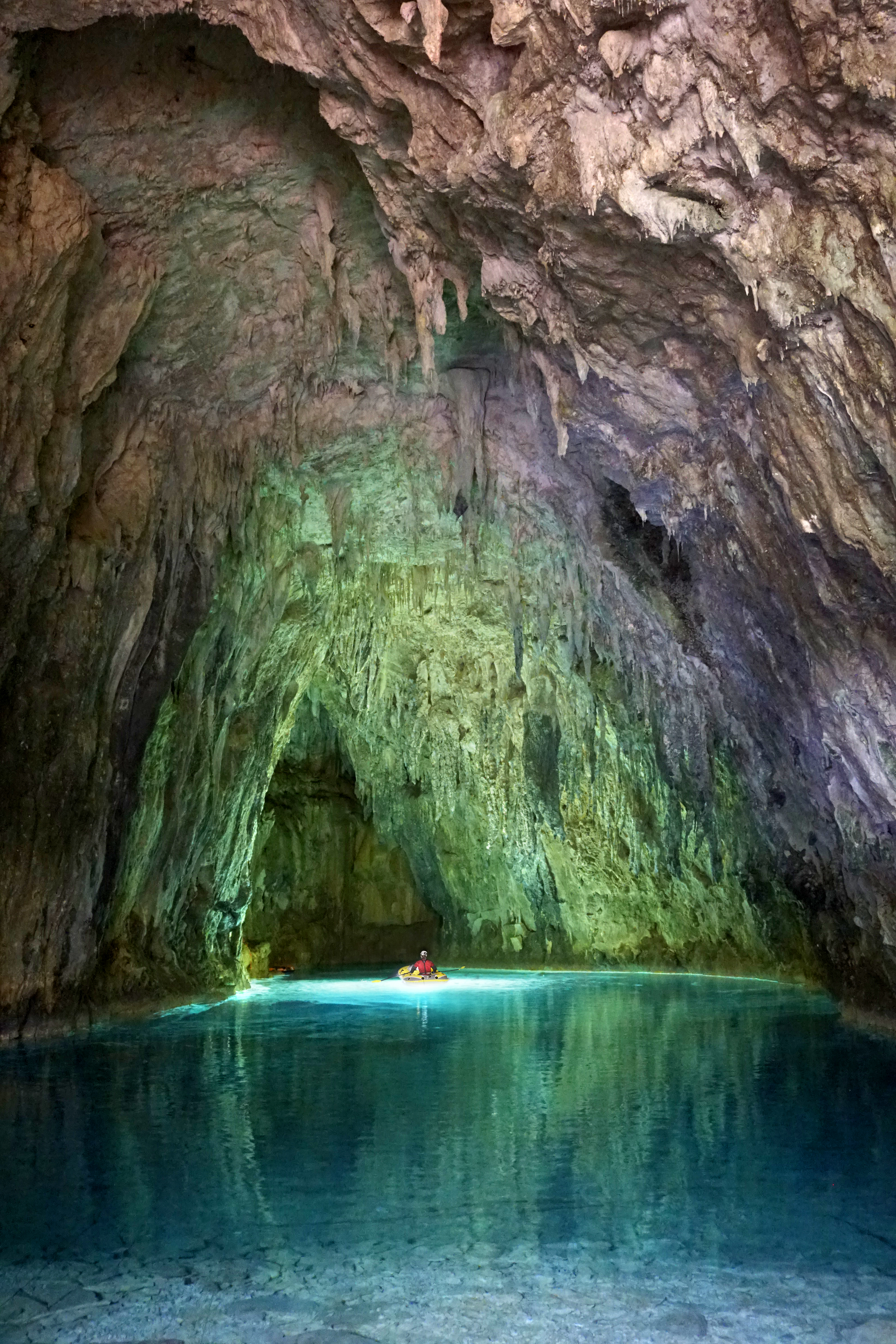 Zoomed out shot of a large cave with light illuminating a tiny person on a boat in the background.