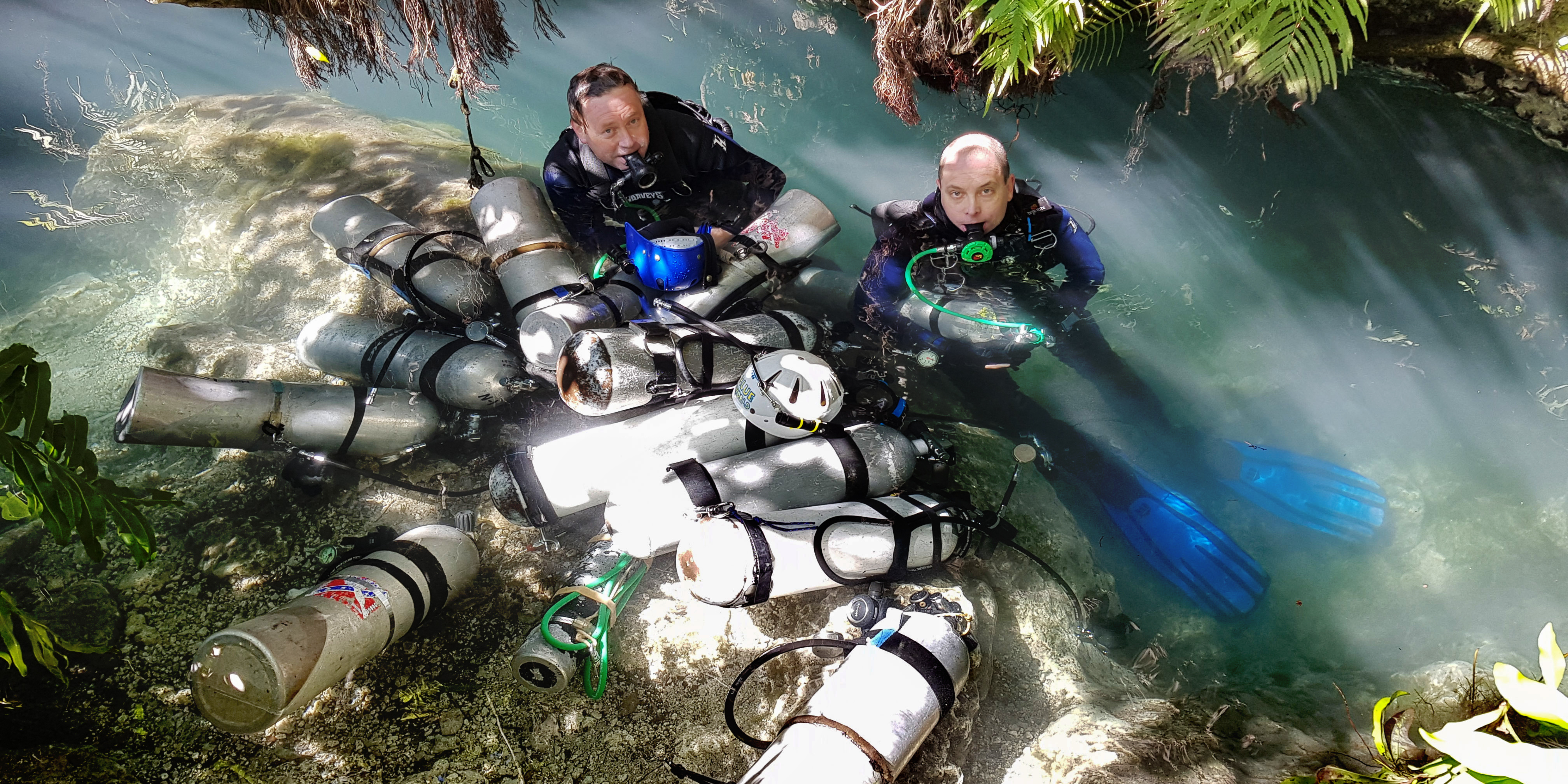 Two people in diving gear emerging from the water surrounded by oxygen tanks in an outdoor environment.