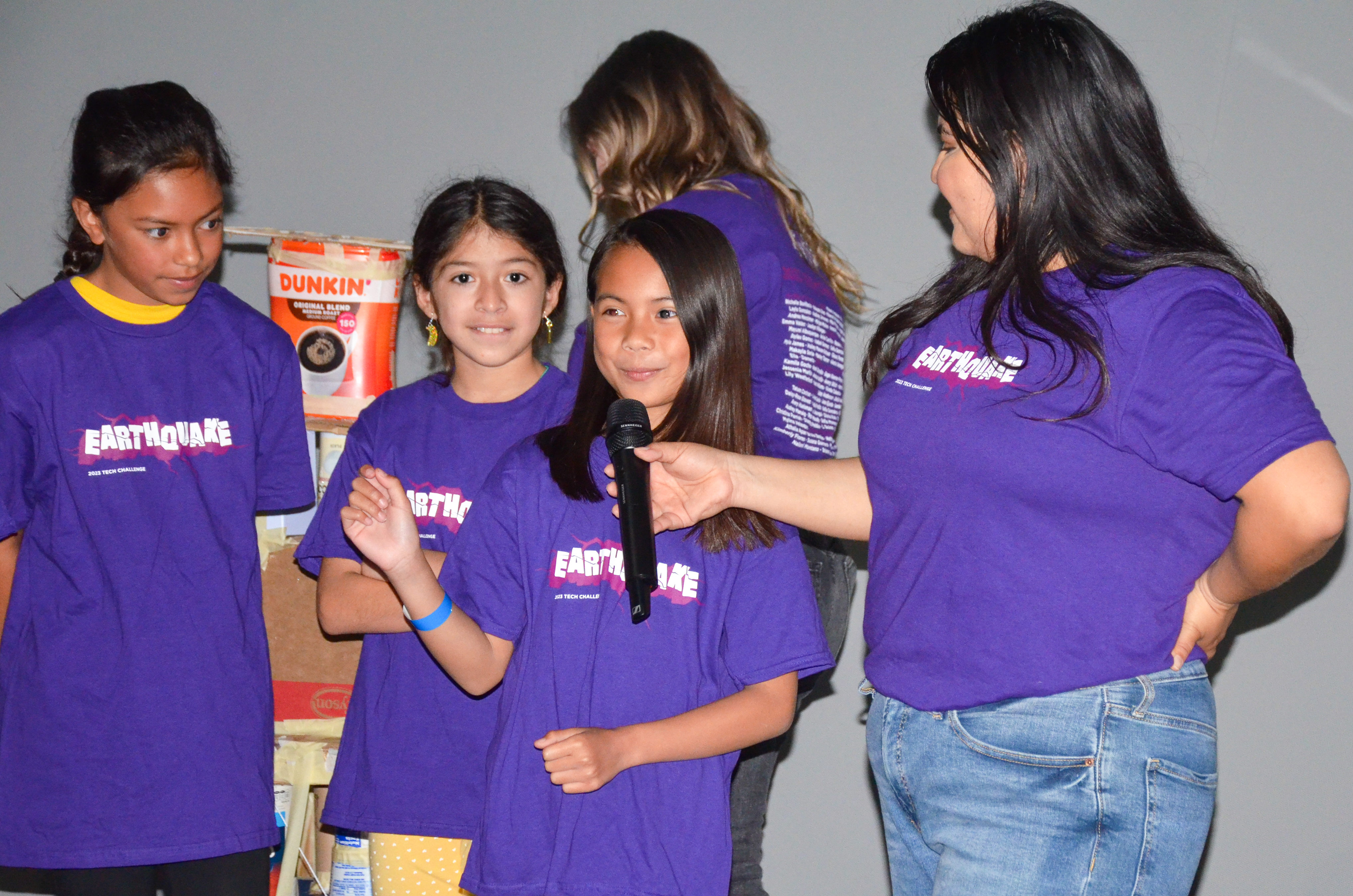 A young woman in a purple t-shirt interviews with a microphone three girls in purple t-shirts while another young woman is in the background. 