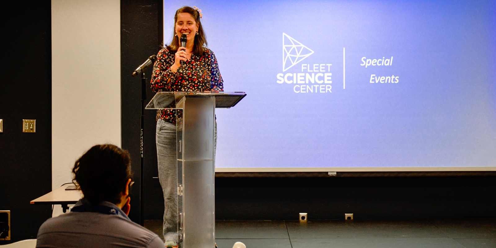 Community Forum A woman stands at a podium in a science center, addressing the audience with enthusiasm.