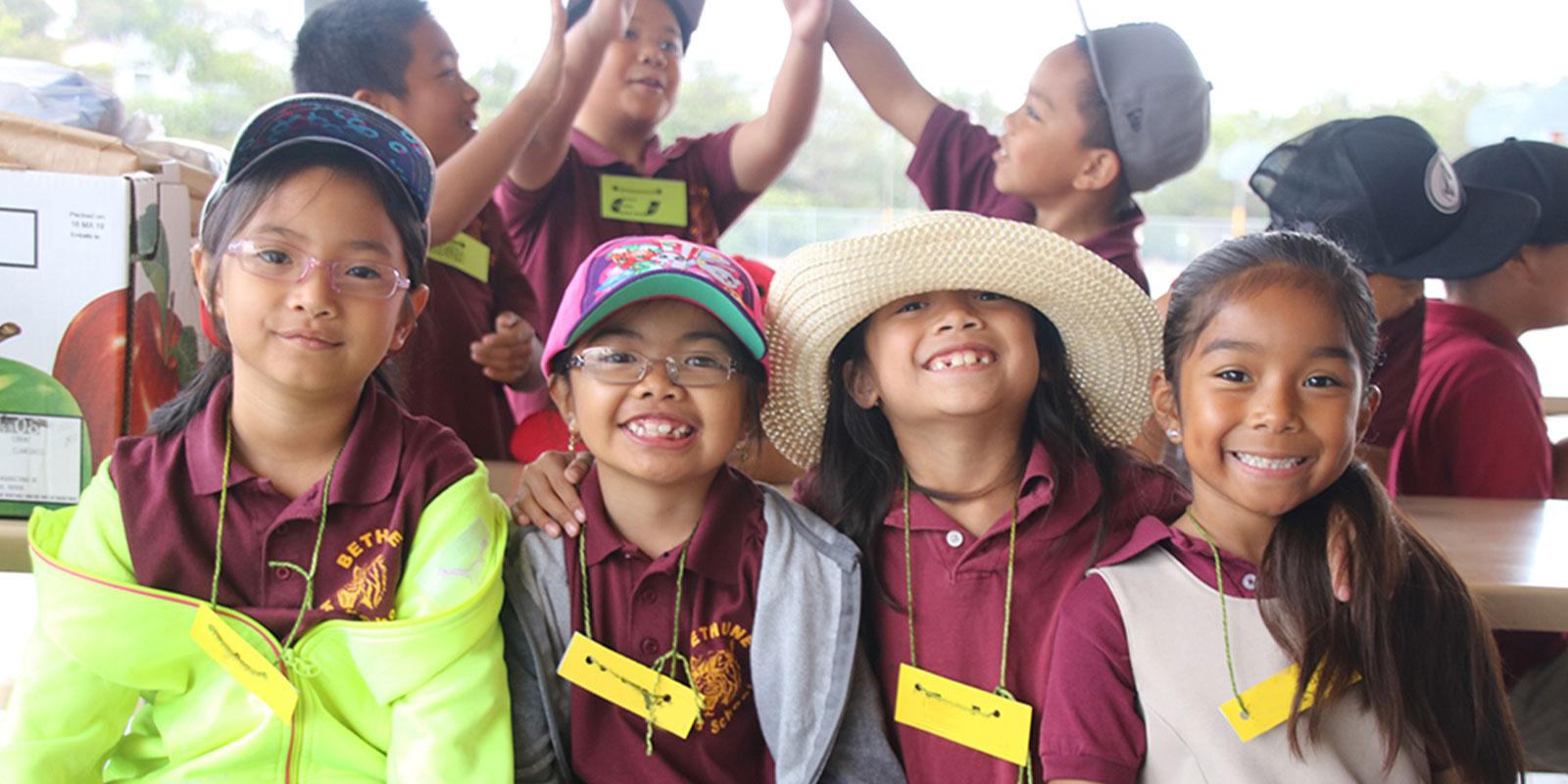 Four elementary school girls in maroon shirts leaning on each other and smiling at the camera