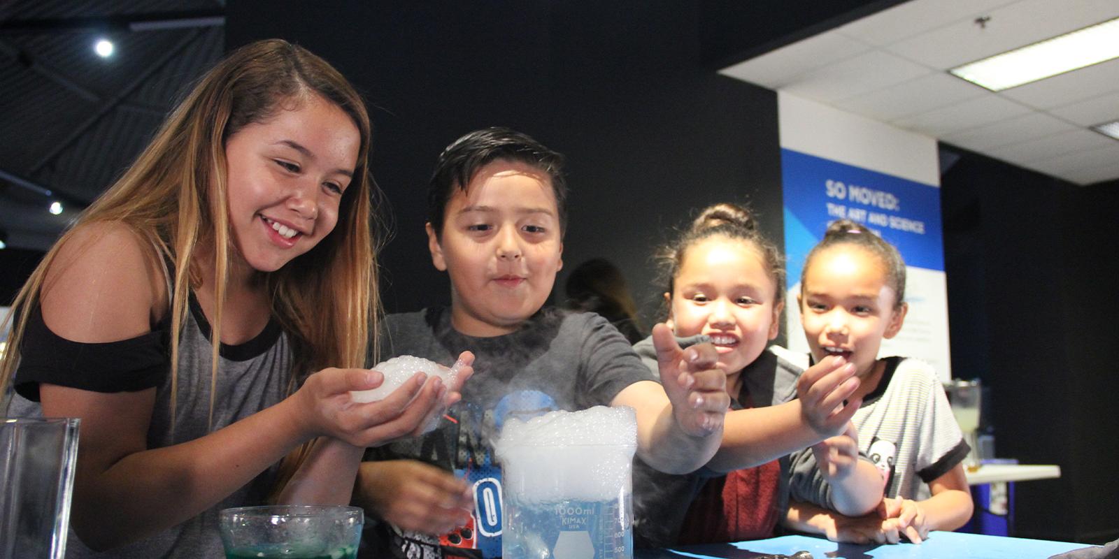 Four school aged kids look on a foaming beaker. The front girl holds foam bubbles in her hand and smiles.