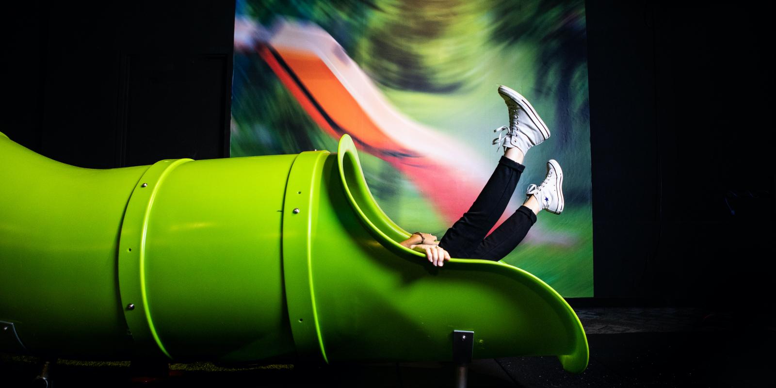 A green slide in a darkened room with a pair of women's legs in jeans coming out the bottom and aimed upward