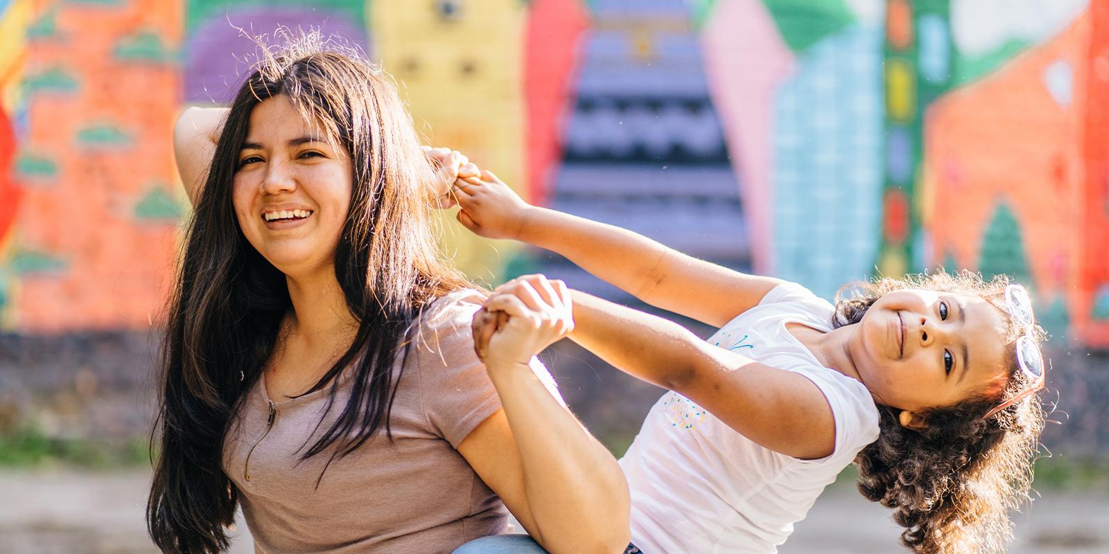 A hispanic woman gives a young hispanic girl a piggy-back ride. They are holding hands and the young girl is leaning back with her head tilted all the way back. They are both smiling and having fun. The background is a vibrant abstract mural. 