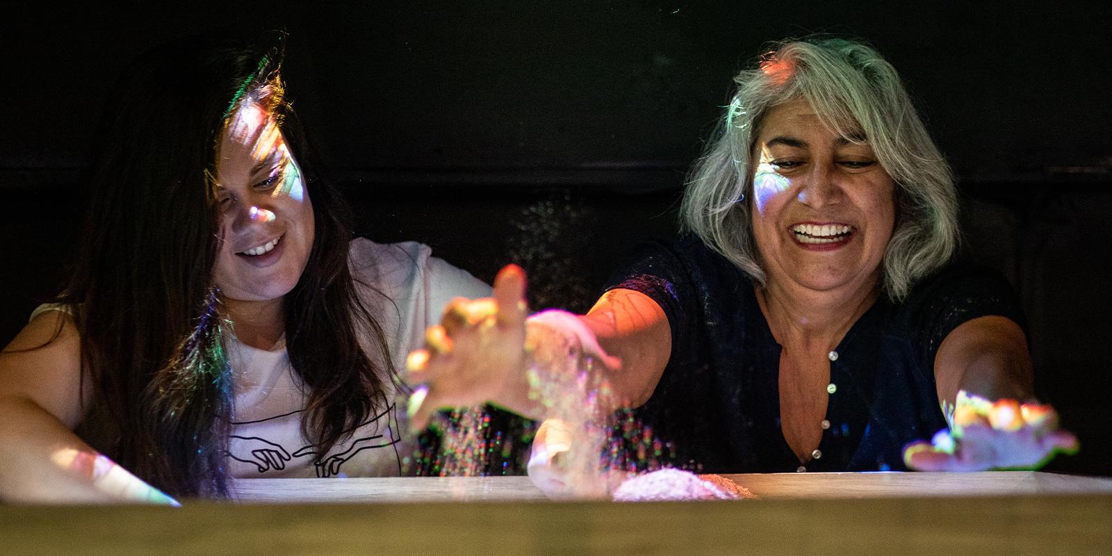 Two women against a black background playing with a kinetic sandbox