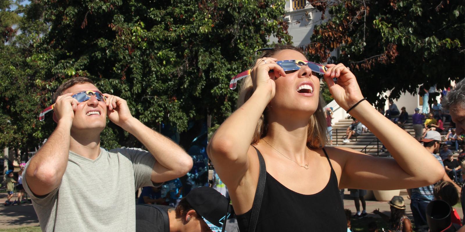 A man and a woman wearing eclipse sunglasses squinting up at an eclipse in a green outdoor area
