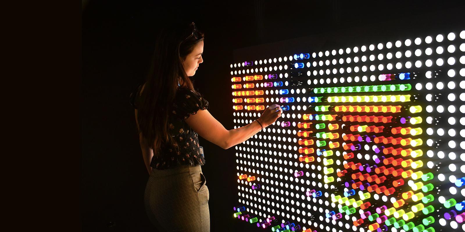 A woman standing in the dark playing with a huge lite brite on a wall