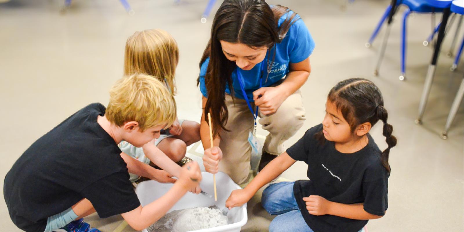 In a bright indoor setting, a fair-skinned, young adult woman with long, dark brown hair and three young children participate in a science project, gathered on the floor around a white plastic container. The woman, likely a science volunteer, assists the children with the experiment, holding a slender wooden rod, possibly a stirring stick, and peering down into the container. She is dressed in a blue short-sleeve shirt featuring a science-related logo and the words "SCIENCE VOLUNTEER," paired with tan khaki