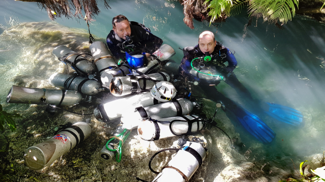 Two people in diving gear emerging from the water surrounded by oxygen tanks in an outdoor environment.