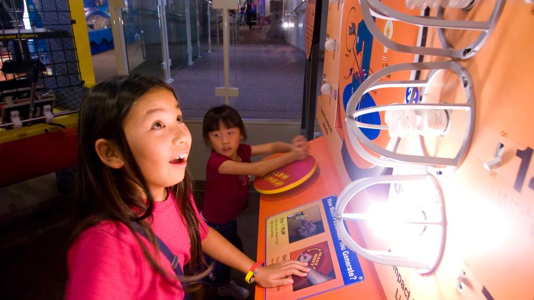 Young dark haired girl looks up with a big smile at a light experiment that's vertical on the wall in front of her at the Fleet Science Center