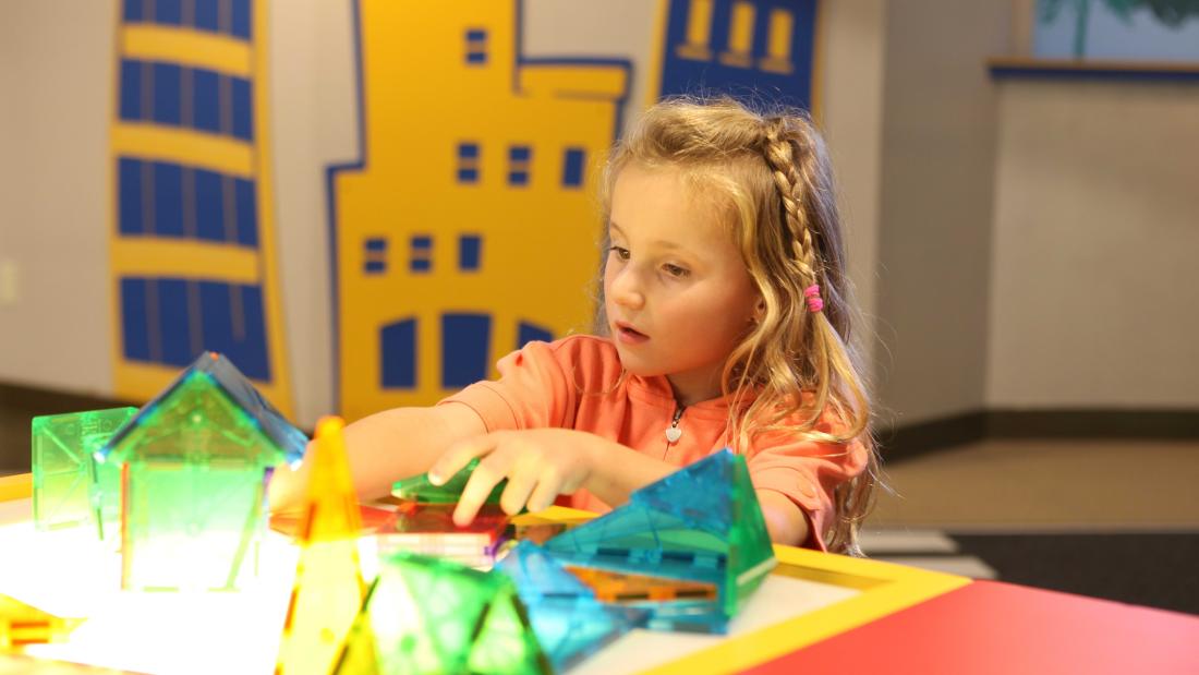 Young blonde girl with a braid plays with colorful clear geometric shapes on a red table at the Fleet Science Center
