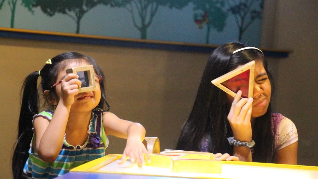 Two young hispanic girls hold colorful shapes up to their eyes and squint through at the Fleet Science Center