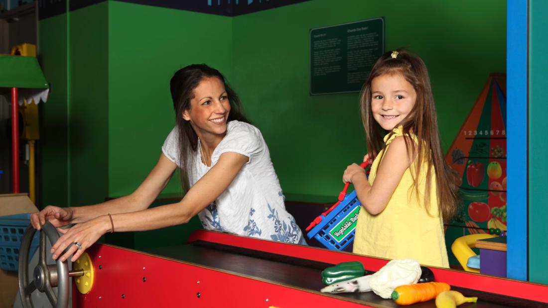An adult brunette woman and a young brunette girl play on a red conveyor belt with plastic fruits and vegetables at the Fleet Science Center
