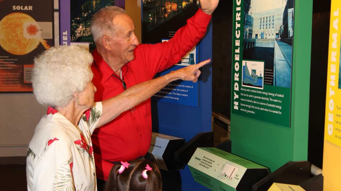 An elderly man and woman point out exhibit information to a young girl in pink with brunette pigtails at the Fleet Science Center