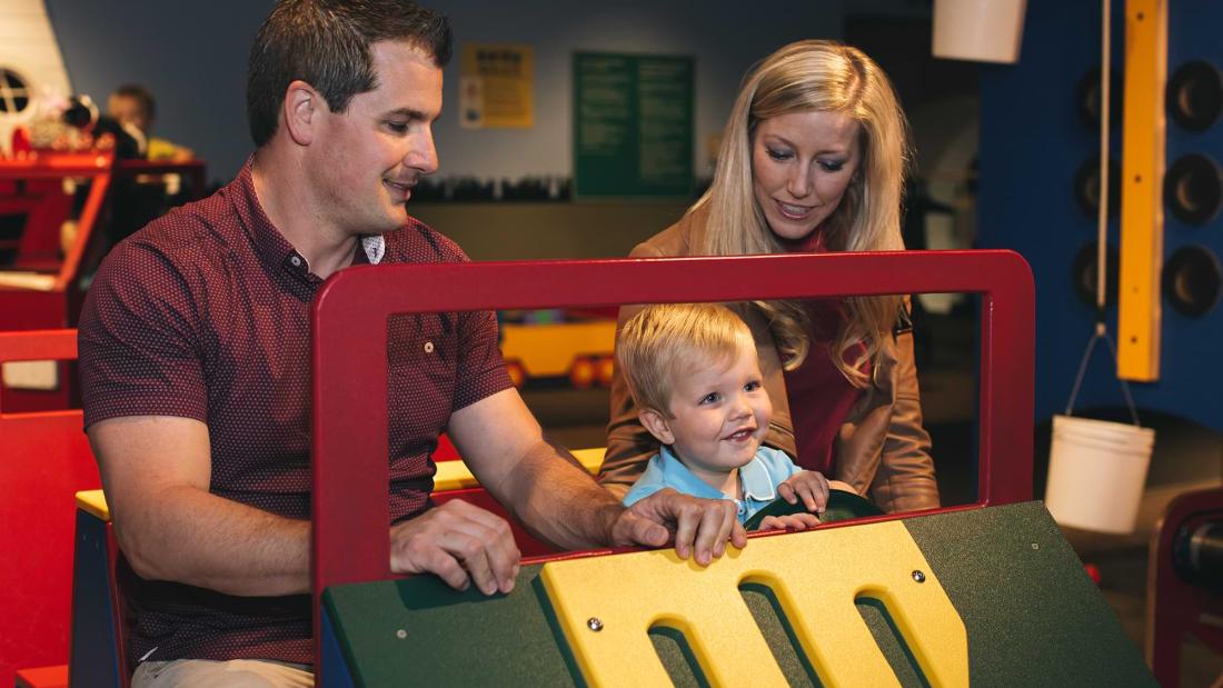 A while man and woman sit in a play car with a blonde child between them at the Fleet Science Center