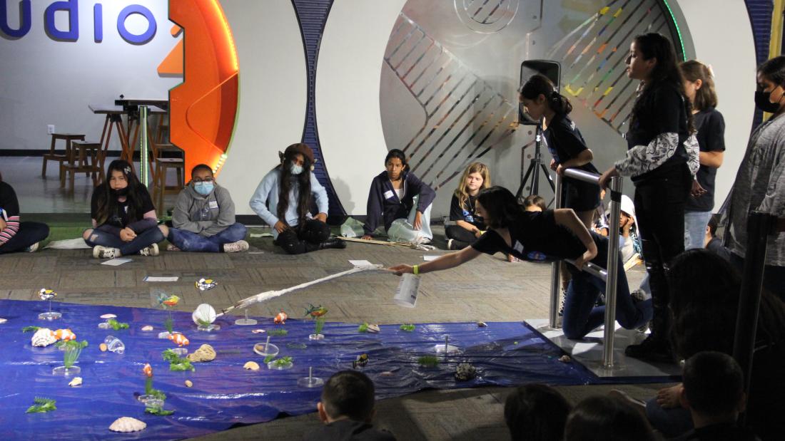 An action shot of girls demonstrating their design. A girl reaches out with crafted equipment into the challenge area which is blue, on the floor, and covered in fake fish and trash.