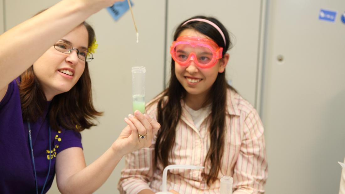 A young girl in safety goggles looks on as a female instructor droppers liquid into a test tube