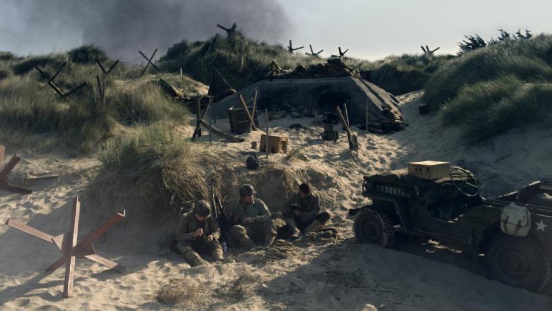 Three men sit on the beach surrounded by military equipment. Smoke is rising in the background.