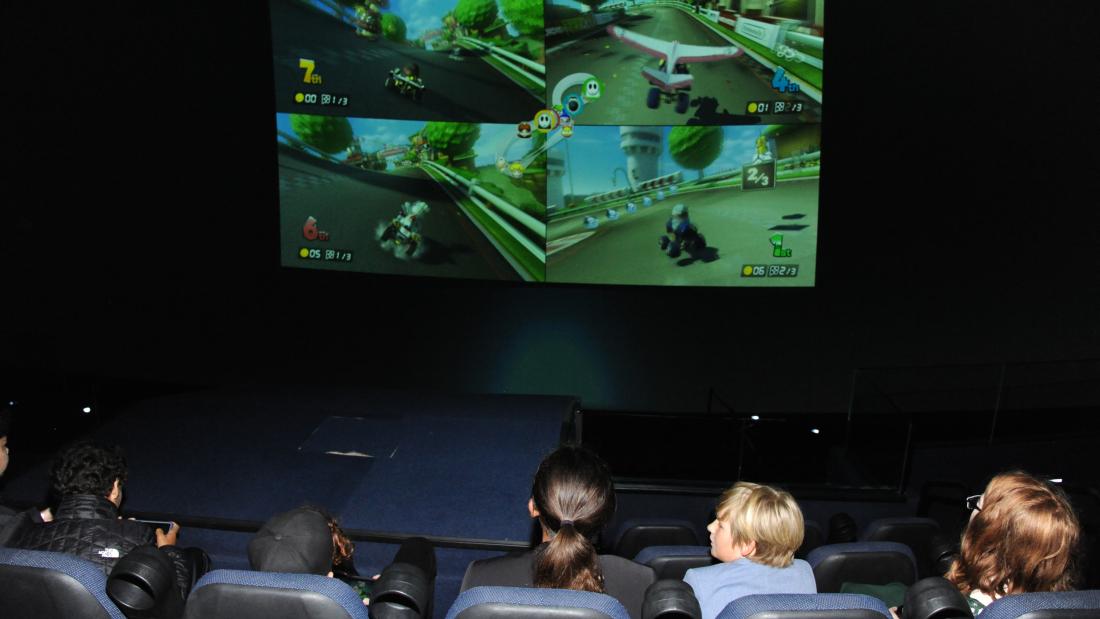 A photo taken from behind a row of young kids sitting in stadium seating. They are playing a four player video game which is projected onto the large imax screen in front of them. 
