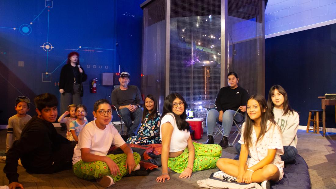 A group of kids in pajamas sit on the floor and smile and make funny faces. A male and female adult are behind them sitting. An adult woman speaks into a microphone while a large tesla coil next to her lights up.