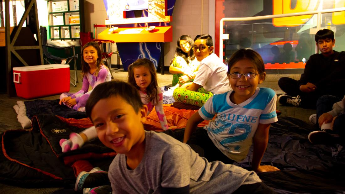 A group of kids in pajamas sit on the floor in sleeping bags and smile at the camera