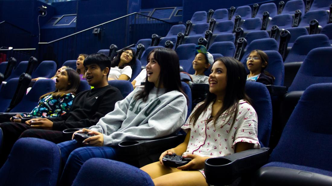 A group of young teens and kids sit in stadium seating with the front three holding game controllers. They are smiling and gasping and laughing while looking at the screen which is out of frame.