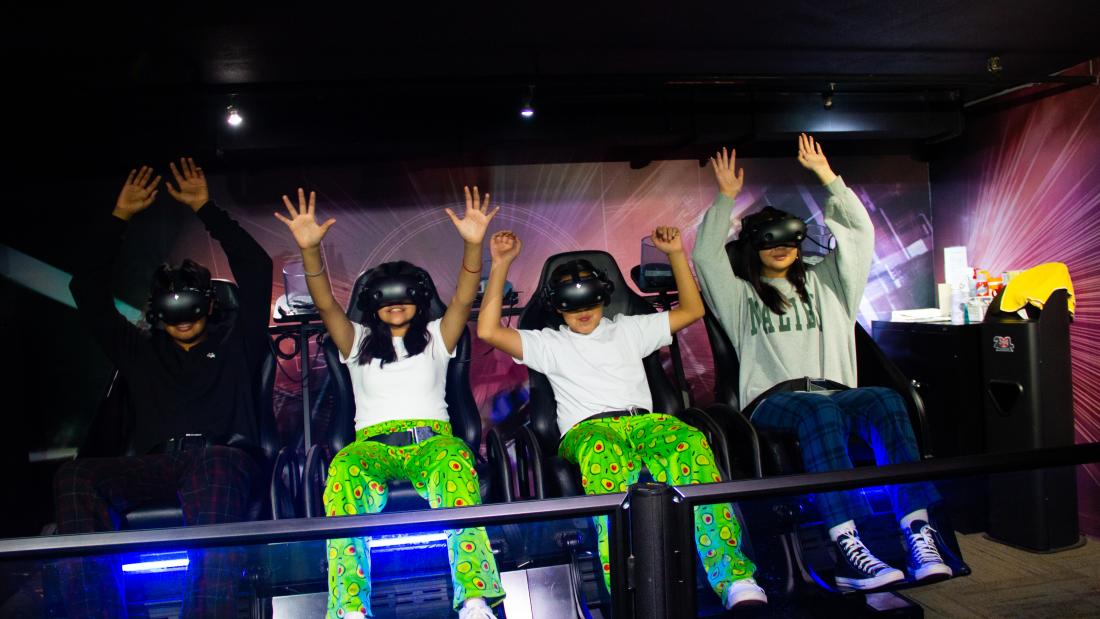 Four kids wear VR headsets and are seated in a VR motion machine. They have their hands up in the air with excitement