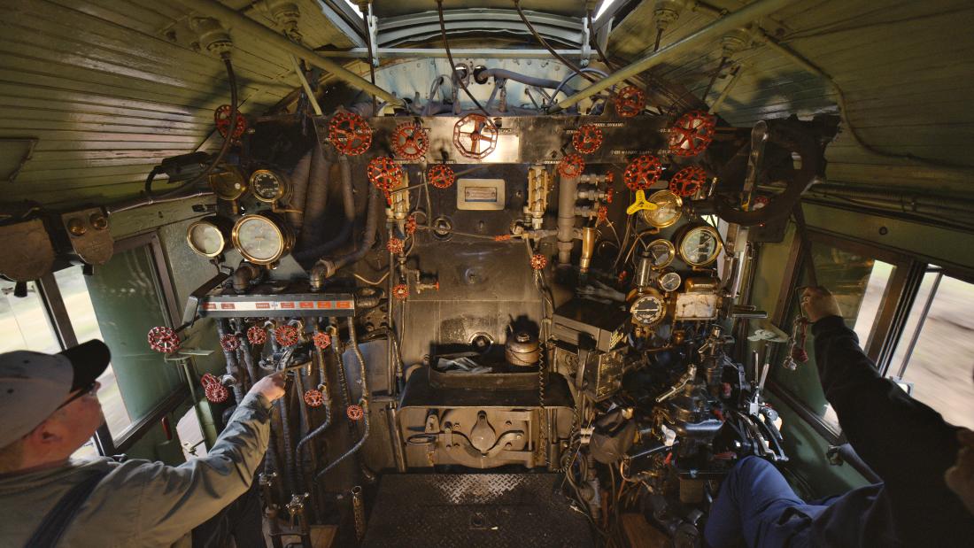 The inside of a train with a conductor standing in the bottom left and reaching for one of the many knows and levers. 