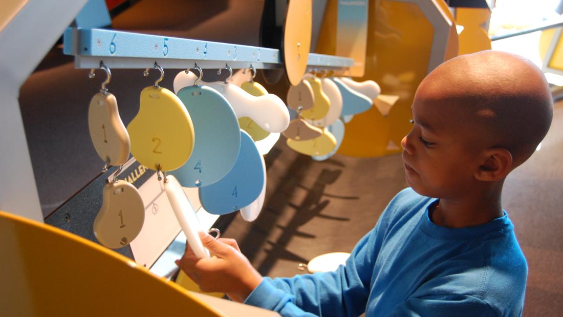 A young black boy holds a hanging oblong object. There are many artistic blob style shapes handing from a blue wooded rod. The shapes are colored white, yellow, and different shades of blue and have numbers written on them. 