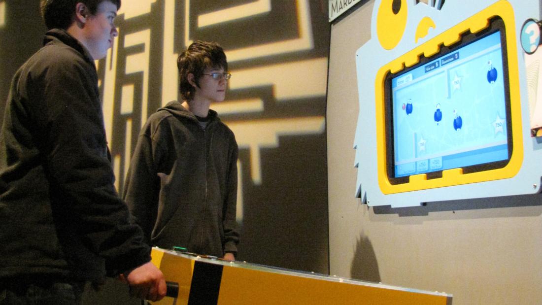 Two white teen boys stand in front a yellow tilt box. They are interacting with a blue screen console that has a fun juvenile monster head shape frame surrounding it. A computer chip pattern is projected onto the wall in light.