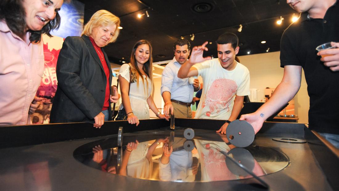 A group of adults stand around a black flat top with a black disc in the center. They are spinning metal discs on the table top disc and look excited. 