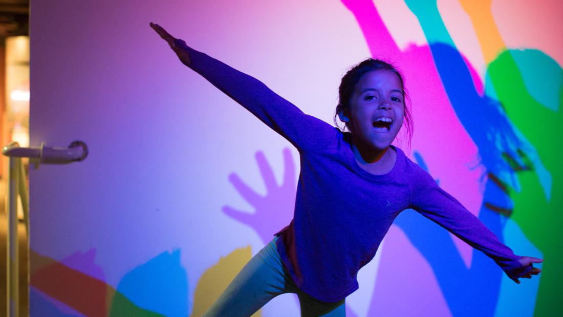 A young girl leans forward with her arms out and mouth open. He shadow behind her is in pink, blue, green, and yellow.