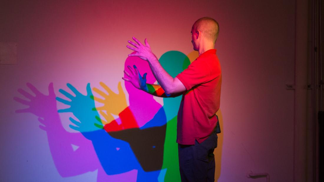 An adult white man in a red shirt faces a wall with his hands out. His shadow is in pink, blue, green, and yellow.