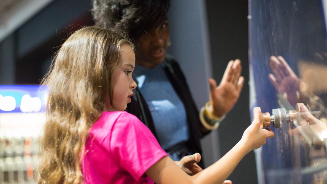 A black woman talks to a young white girl who is pulling a knob on an exhibit piece 
