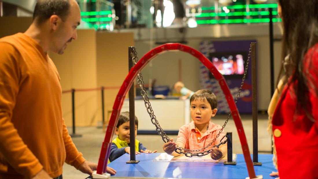 An adult man on the right, two small male children in the middle, and an adult woman to the right. There is a standing red arch of blocks in the center that the children are working on building.