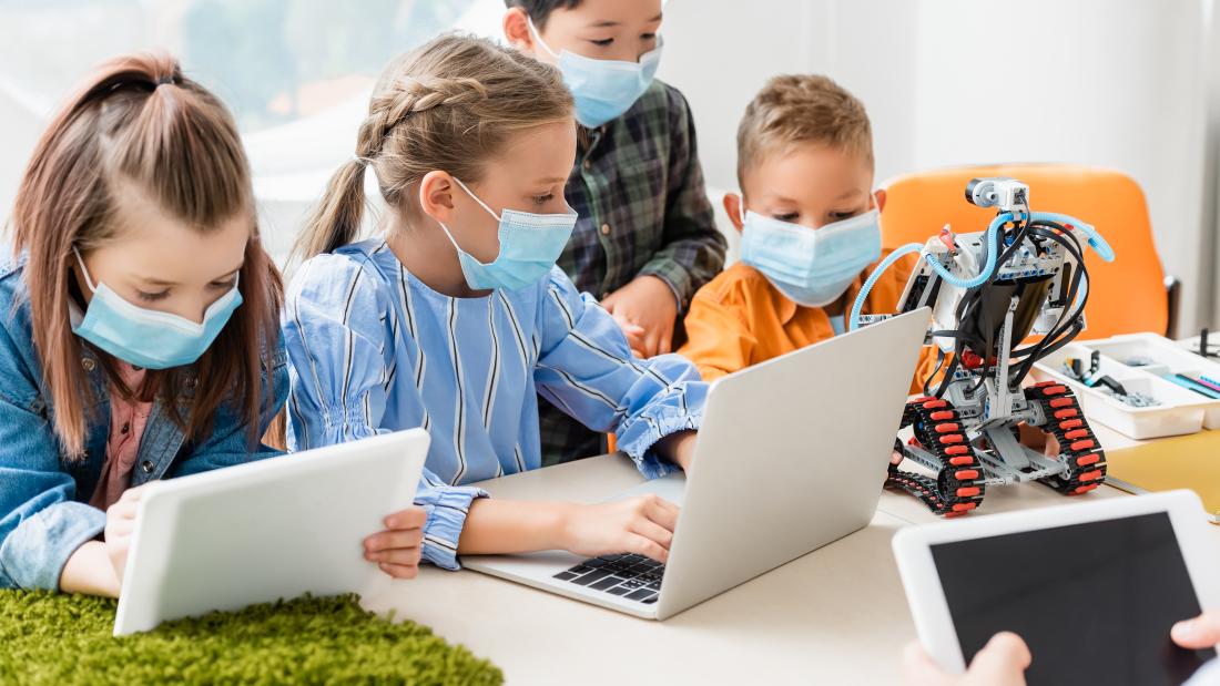 Four students wearing masks and using various forms of handheld and computer technology in a classroom.