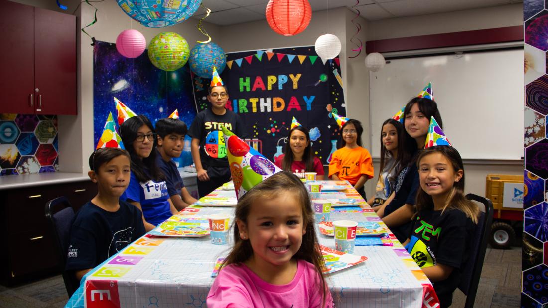 A group photo of children with birthday hats sitting around a long table. Festive decor hangs from the ceiling and the wall says Happy Birthday.