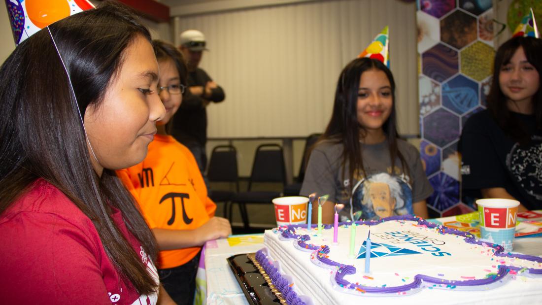 A close up profile of a birthday girl with a lit cake in front of her while three girls smile along the sides of the table.