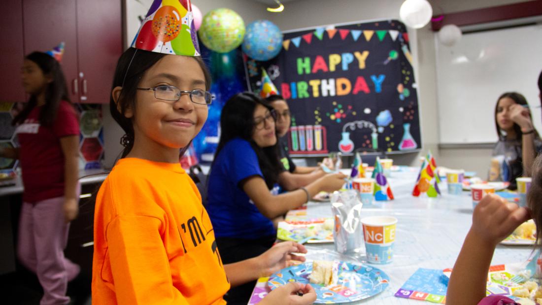 A girl in an orange shirt and birthday hat smiles at the camera with cake on her plate and fork.